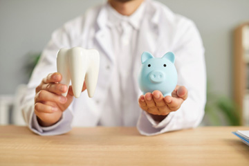 Dentist holding model tooth in one hand and blue piggy bank in the other
